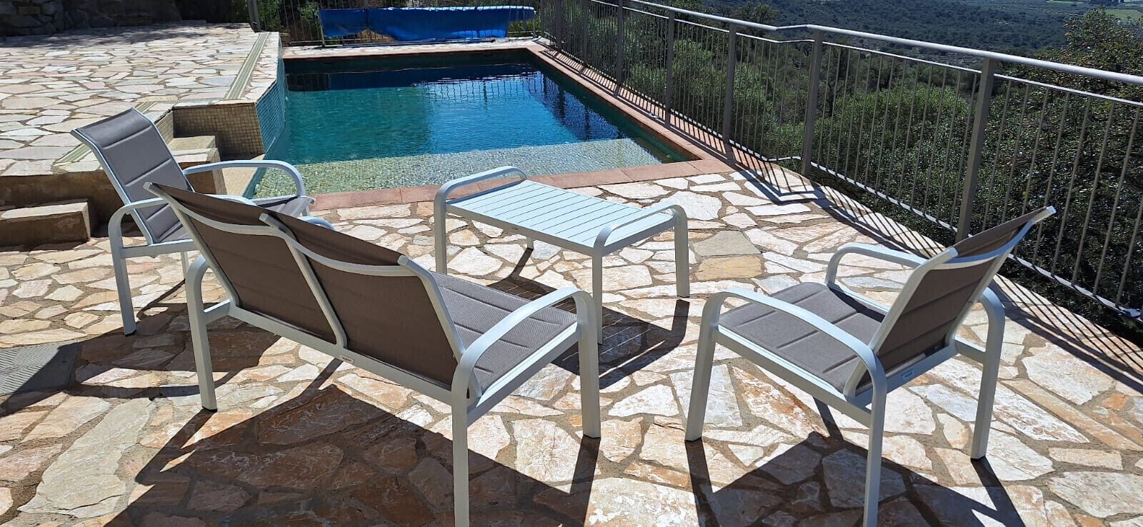 Zona junto a la piscina con suelo de piedra, piscina y mesa con sillas.