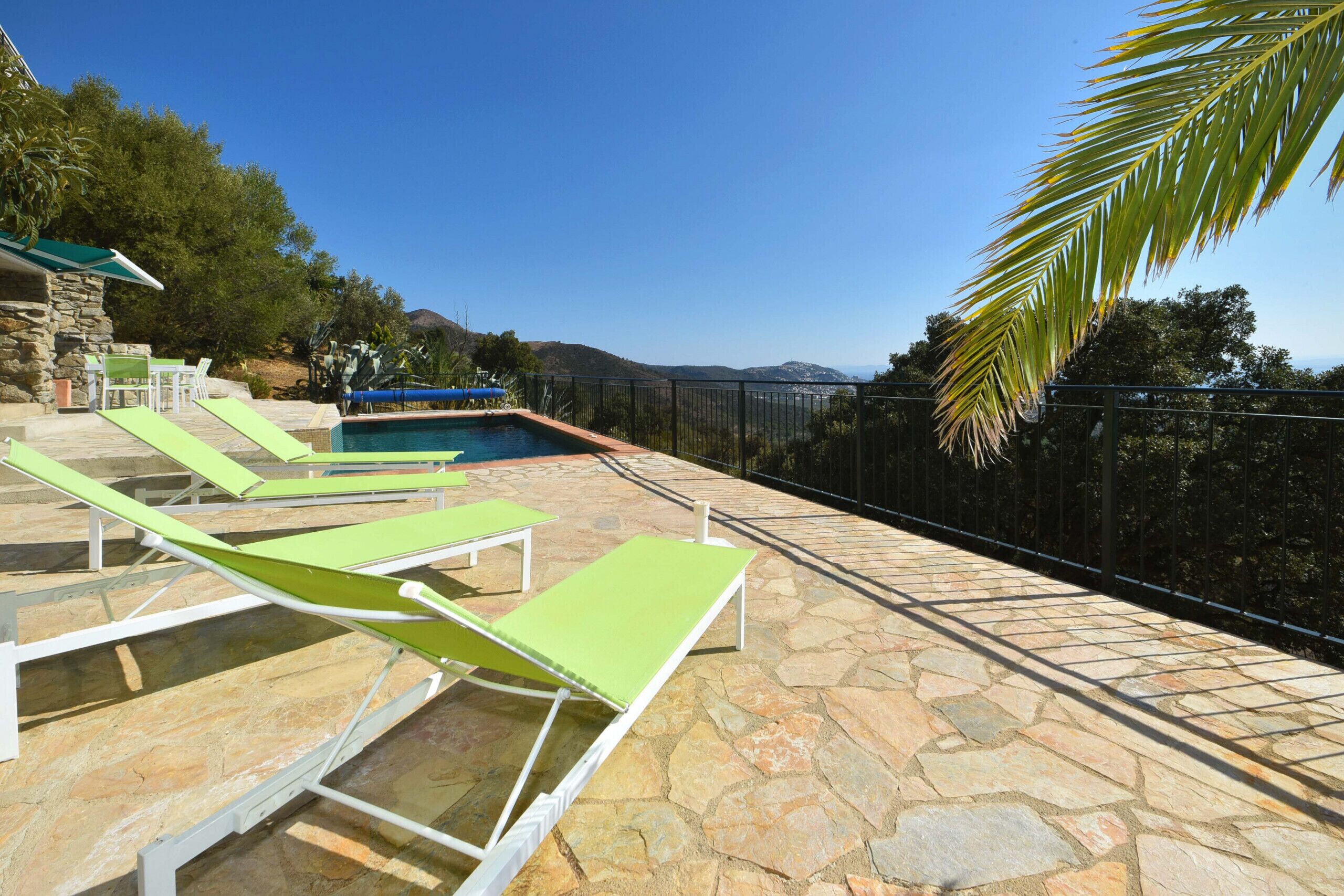 Zona de piscina con suelo de piedra, sillones reclinables verdes y vistas al mar.