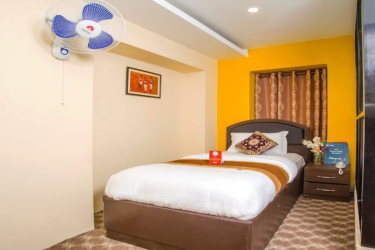 Een hotelkamer met een bed, nachtkastje, ventilator, een ingelijste foto en een raam met gordijnen.