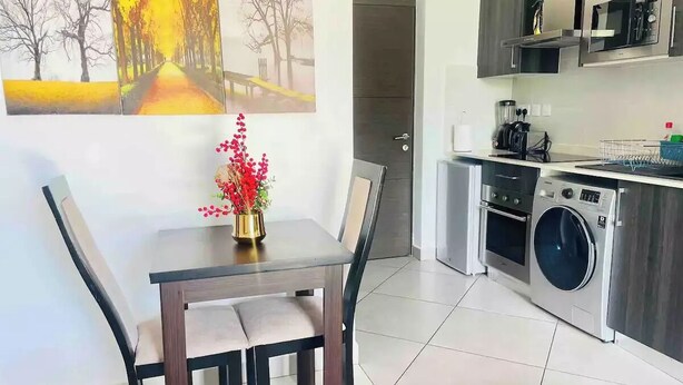 Une cuisine moderne équipée d’un four encastré, d’un four à micro-ondes et d’un lave-linge. On y trouve une table à manger avec des chaises et un vase de fleurs.
