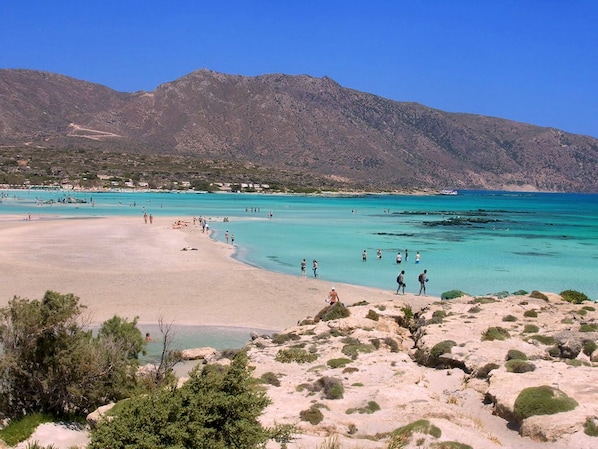 A beach with clear turquoise waters, rocky terrain, and mountains in the background.