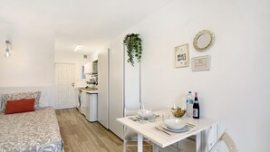 Una habitación moderna y bien iluminada con una cama, una mesa de comedor para dos personas y una cocina al fondo.