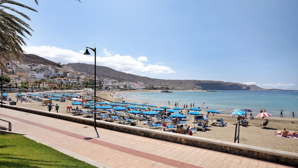 Una playa con tumbonas y sombrillas, un paseo marítimo empedrado y edificios al fondo.
