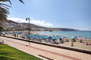 Una playa con tumbonas y sombrillas, un paseo marítimo empedrado y edificios al fondo.