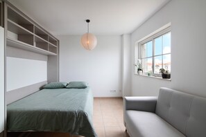 Une chambre avec un lit, un canapé gris, une fenêtre donnant sur un beau paysage et un luminaire suspendu.