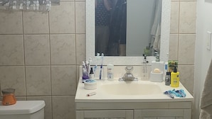 A bathroom with a white sink, mirror, and tiled walls. There is a toilet to the left and a small shelf under the sink.