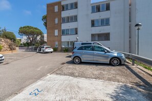 Un parking avec un bâtiment blanc, un arbre et une voiture argentée garée.