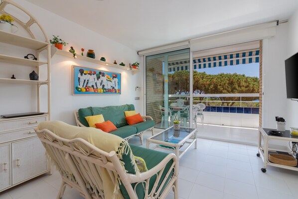 Un balcon avec une porte vitrée, un canapé vert avec des coussins orange, une chaise en osier, une armoire blanche et un tableau accroché au mur.