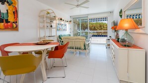 A bright room with a dining table, chairs, a crib, a bookshelf, and a large window. - Apartment 'Les Magnolies' with Sea Views, Wi-Fi and Air Conditioning (PLAYA  DE  ARO)