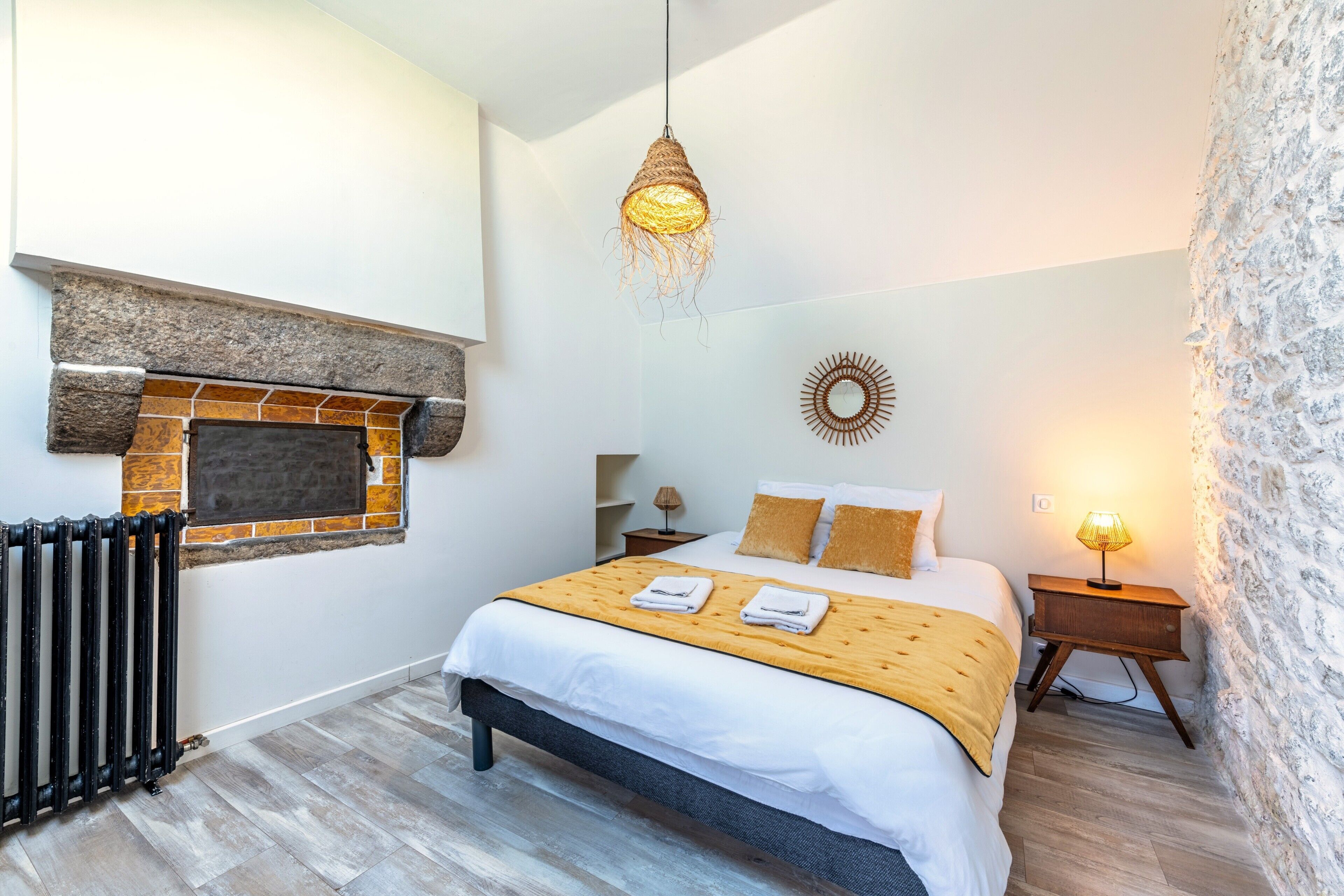 A bedroom with a stone fireplace, a bed with a yellow blanket, a hanging light fixture, and a small bedside table with a lamp.