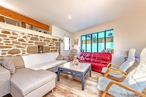 A living room with a stone wall, a red sofa, a wooden coffee table, and a large window. - Holiday Home 'Villa Léonie – Indoor Pool' with Terrace, Garden, and Wi-Fi (Kerlouan)
