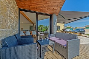 Une terrasse extérieure avec des meubles en osier, une table avec des boissons et une vue sur l’intérieur à travers des portes vitrées.