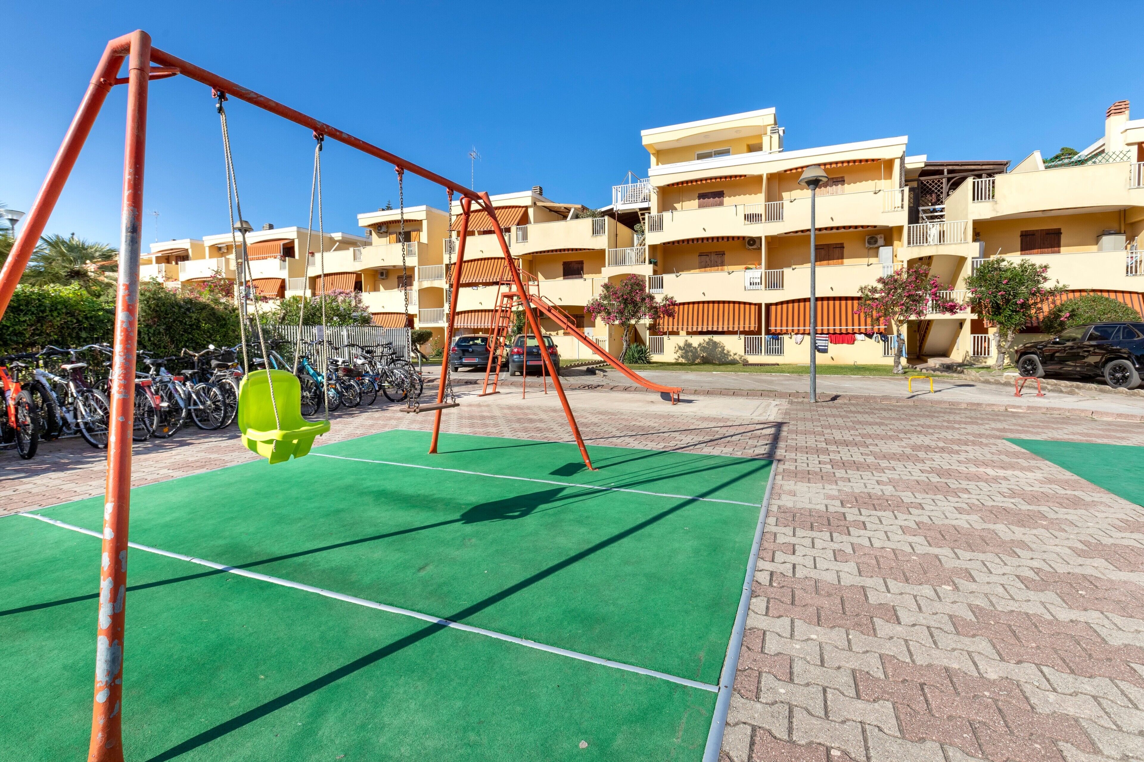 Un parque infantil con columpios y suelo verde, rodeado de bicicletas estacionadas y una hilera de edificios amarillos con balcones.