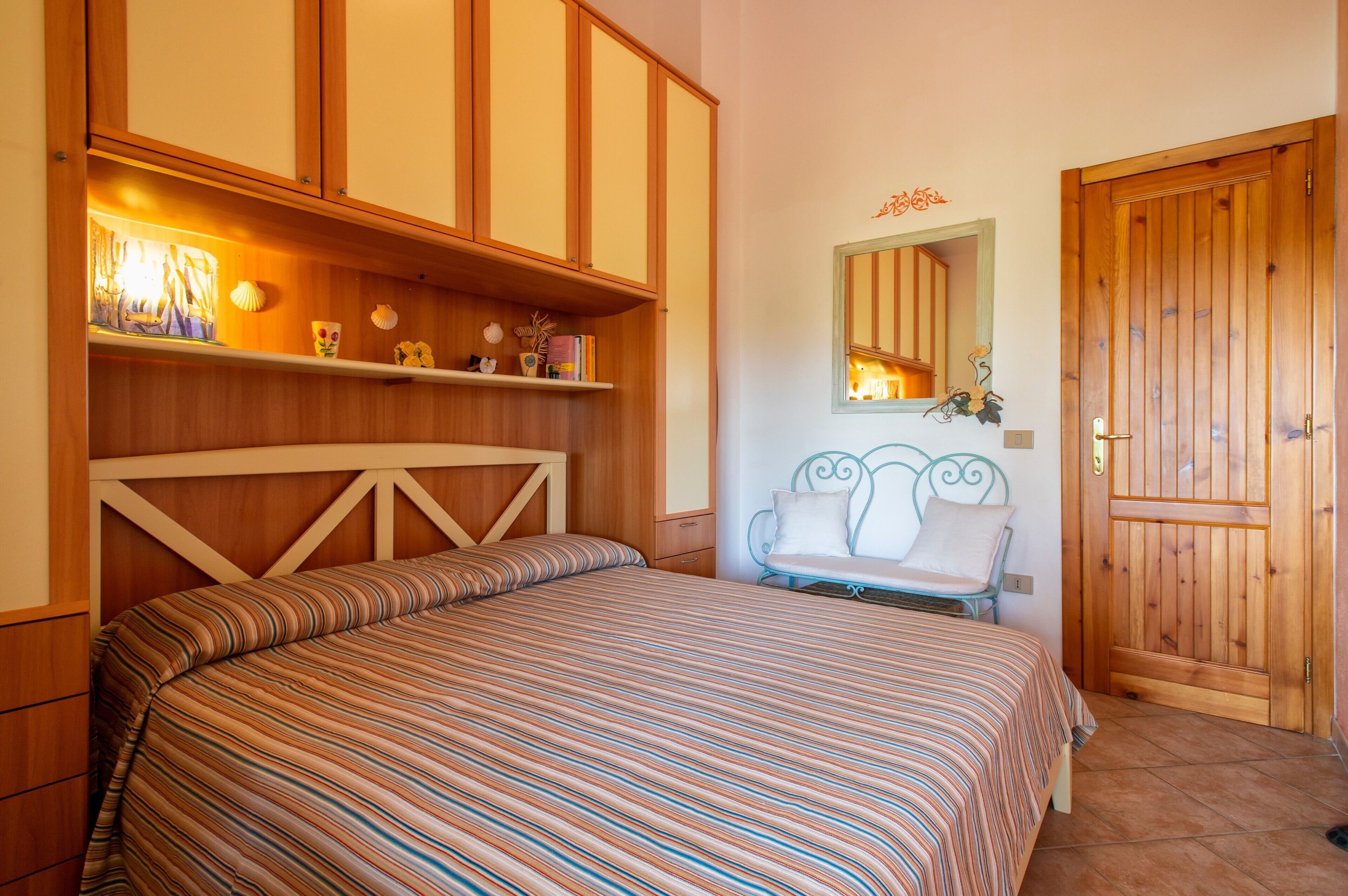 A bedroom with a bed, wooden wardrobe, shelf with decorative items, a mirror, and a door.