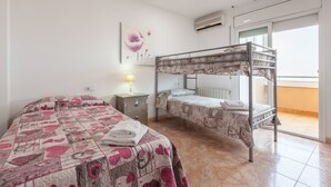 Un dormitorio con una cama litera, una cama individual, una mesita de noche con lámpara, aire acondicionado instalado en la pared y una ventana con vistas.
