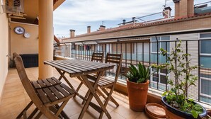 Un balcón con mesa y sillas de madera, plantas en macetas y vistas a edificios vecinos.