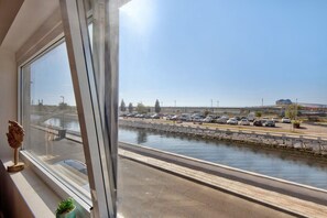 Une vue depuis une fenêtre donnant sur un parking et un canal, avec une petite plante posée sur le rebord de la fenêtre.