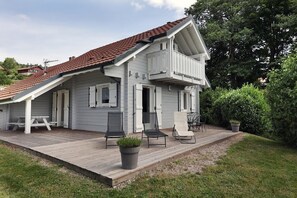 Una casa de dos plantas con tejado de tejas rojas, un balcón y una terraza de madera con mobiliario exterior.