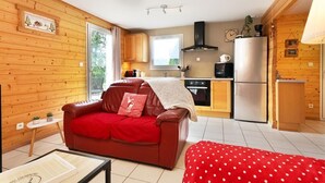 Un salon chaleureux avec un canapé rouge, une table basse et un plaid à pois. On aperçoit, au fond, une cuisine avec des meubles en bois et un réfrigérateur en acier inoxydable.