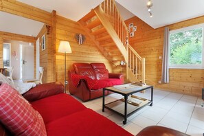Interior de una cabaña de troncos con una escalera de madera, un sofá de cuero rojo, un sillón blanco y una mesa de comedor.