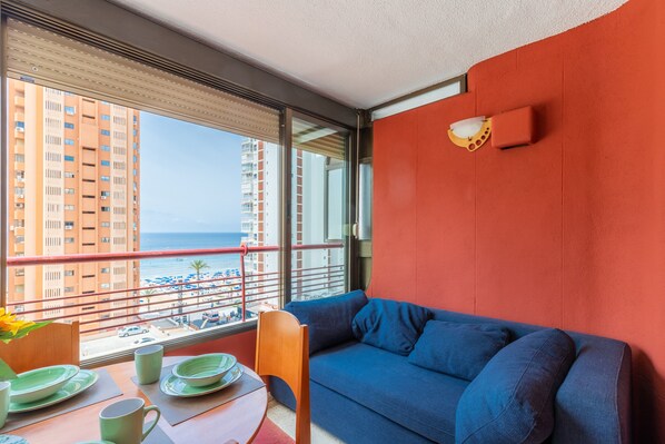 A balcony with a view of the sea and buildings, a blue sofa, and a table with plates and a vase.