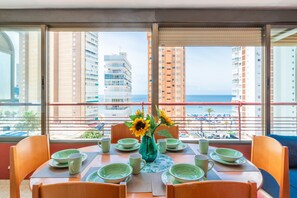 A dining area with a table set for four, a vase with sunflowers, and a view of a beach and high-rise buildings.