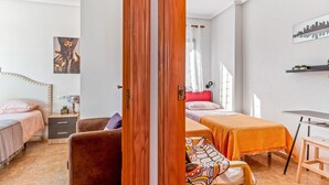 Quarto com duas camas, uma escrivaninha, uma cadeira, mesa de cabeceira e uma porta.