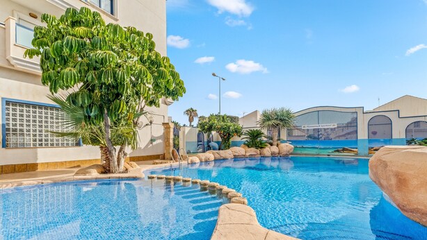 Uma piscina com água azul cristalina, rodeada por pedras e plantas, e um edifício com o letreiro 'Resort Apartments'.