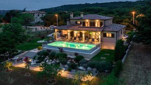 A two-story house with a swimming pool, surrounded by a landscaped garden and hills in the background.