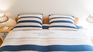 Un lit avec une literie à rayures et une tête de lit, une table de chevet avec une lampe, et une enseigne sur laquelle on peut lire : « RIVIERA MAISON AMSTERDAM ».