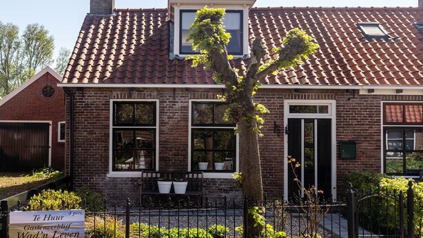 Une maison en briques avec un toit en tuiles, un porche à l’avant orné de plantes en pot et une enseigne devant l’entrée.