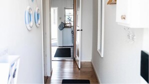 Un couloir menant à une cuisine avec des meubles blancs, un paillasson noir et un porte-manteau suspendu.
