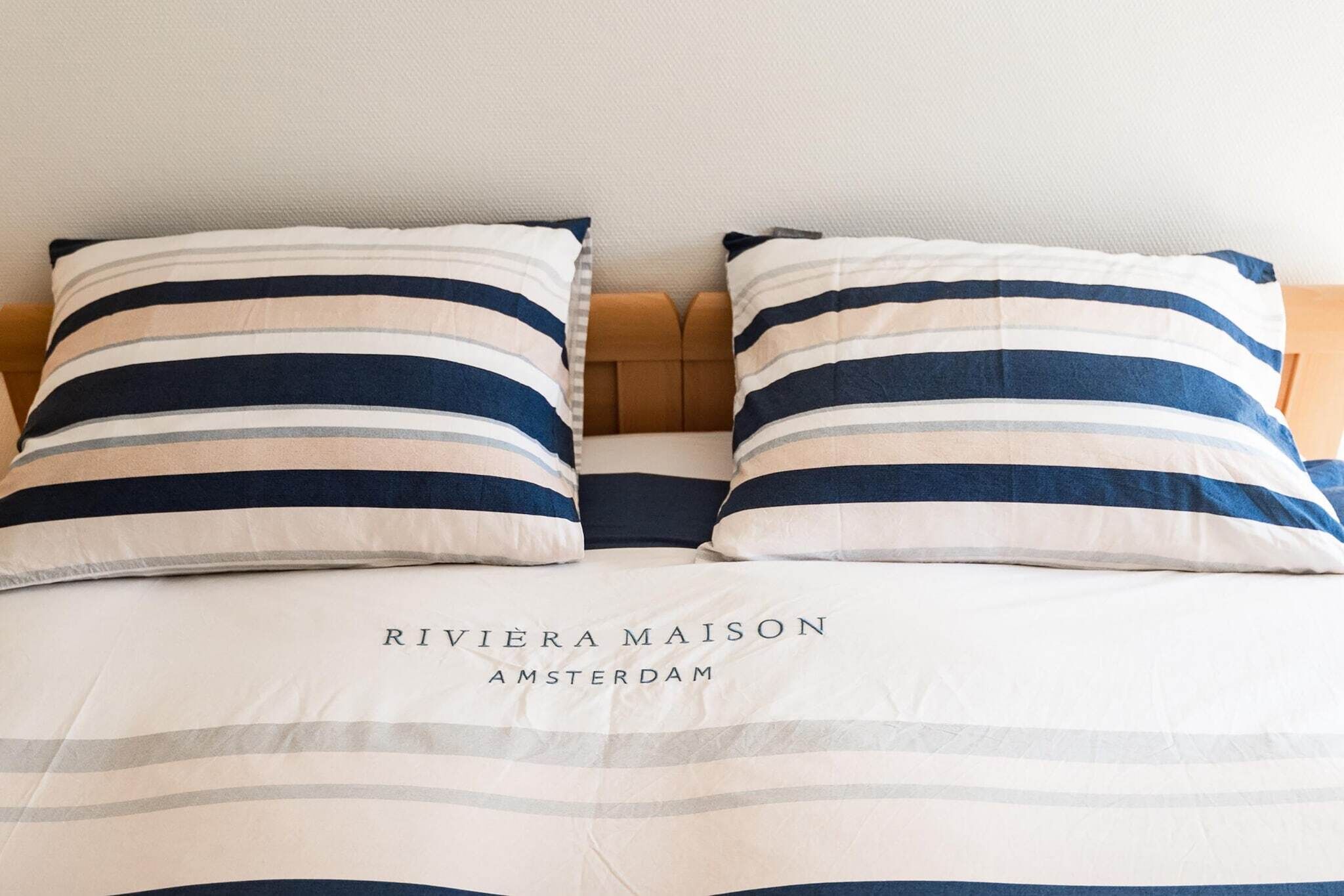 A bed with striped pillows and a headboard, and the text "RIVIERA MAISON AMSTERDAM" on the bedspread.