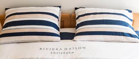 A bed with striped pillows and a headboard, and the text "RIVIERA MAISON AMSTERDAM" on the bedspread.