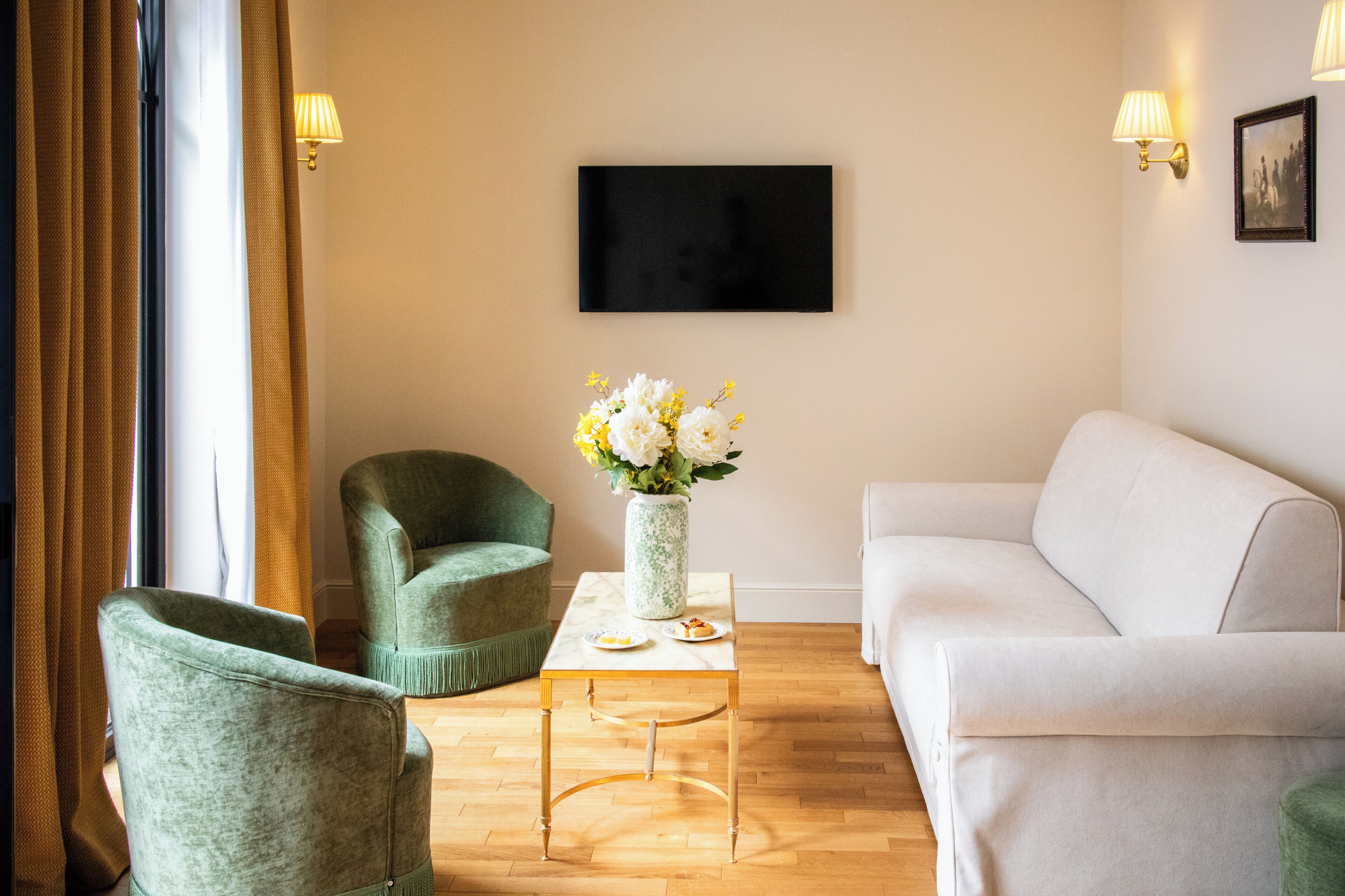 A living room with a sofa, two armchairs, a coffee table, a vase with flowers, a flat-screen TV, and wall-mounted lamps.