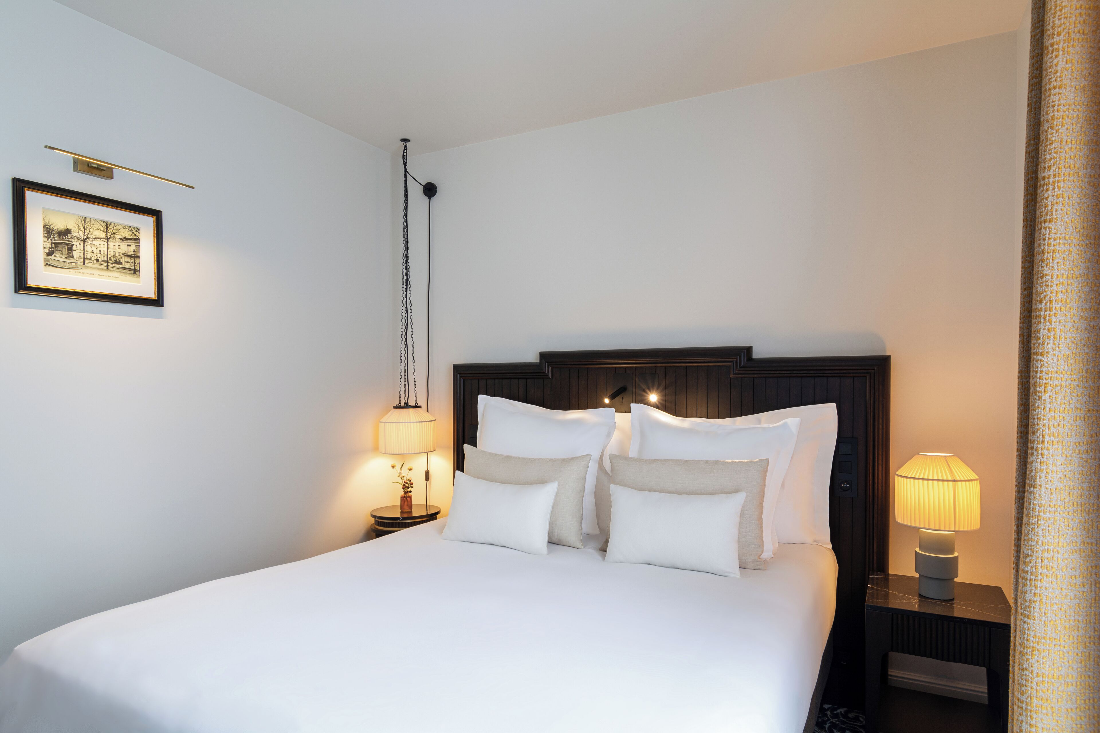 Un lit bien fait, avec du linge de lit blanc et plusieurs oreillers, une table de chevet avec une lampe et un tableau encadré au mur.