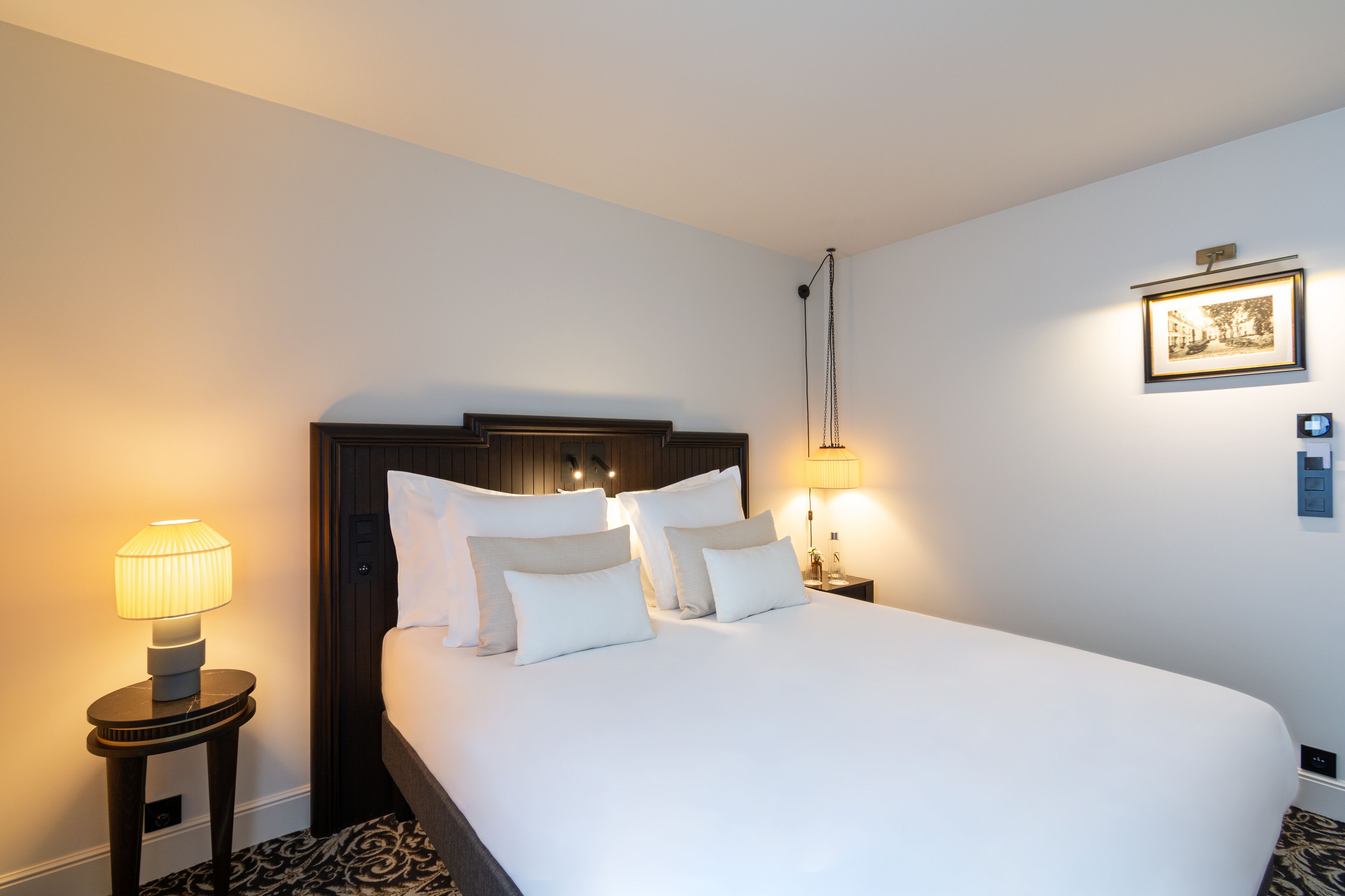 Habitación de hotel con una cama grande, dos lámparas de noche, un cuadro enmarcado en la pared y una mesita con una lámpara.