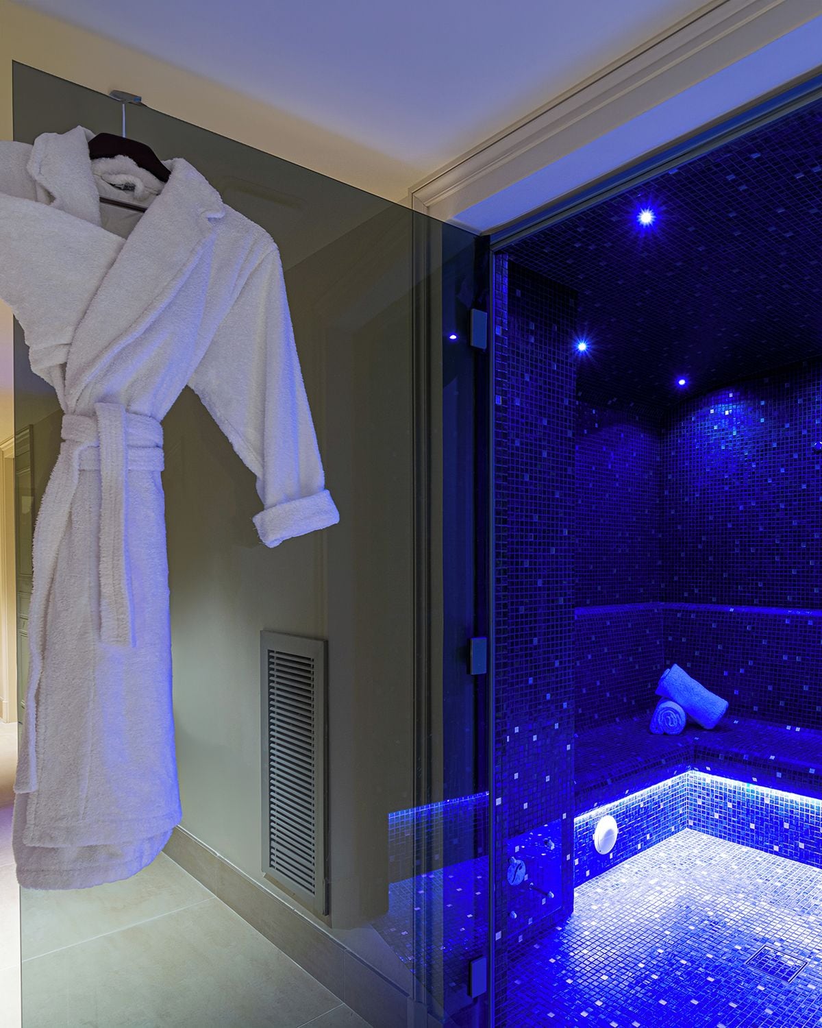 A glass-enclosed room with a blue-lit floor and a white bathrobe hanging on the wall.