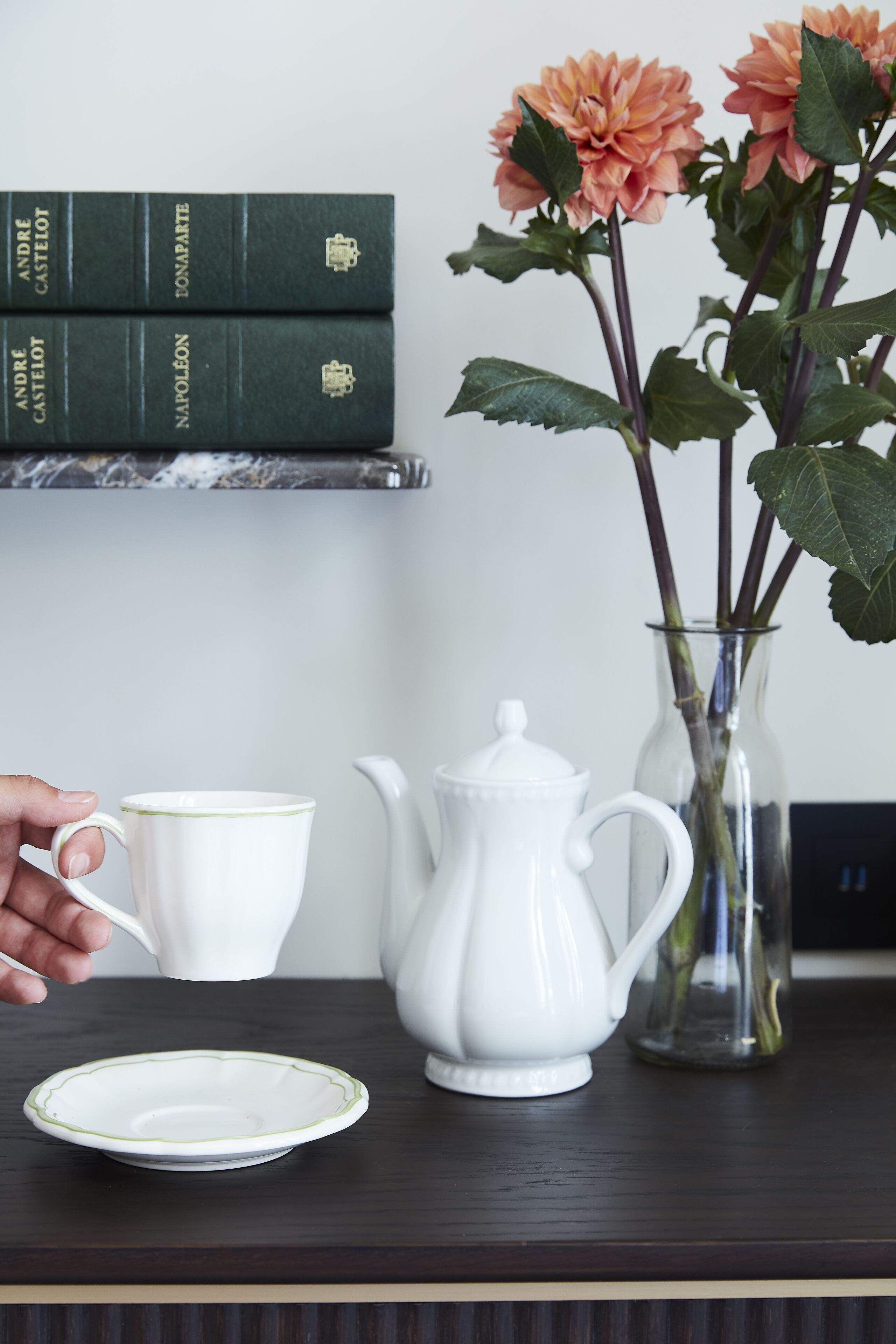 A white teapot, a white cup, a white plate, a vase with flowers, and a stack of books on a dark surface.