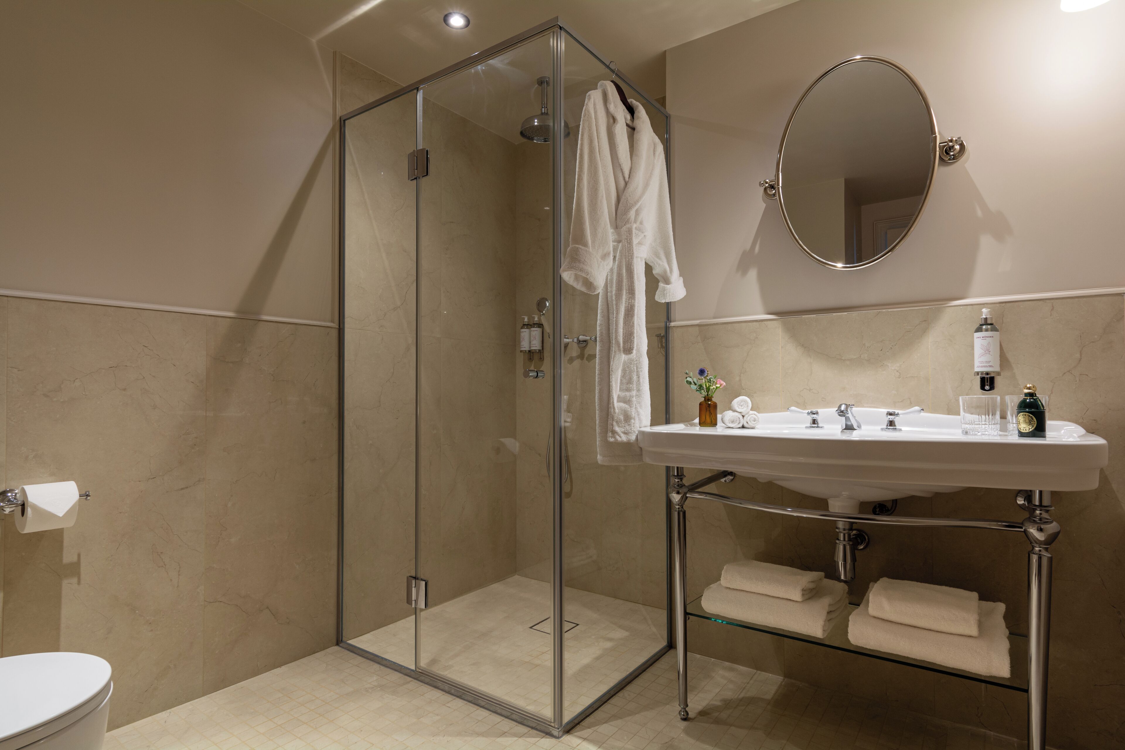 Un baño moderno con ducha de vidrio, un lavabo blanco con espejo y toallas ordenadas en un estante.