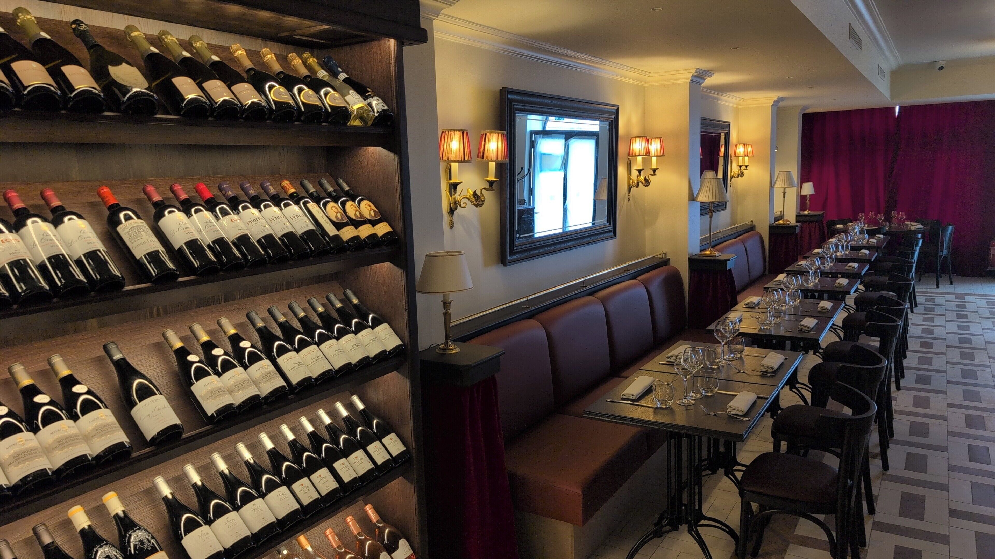 Une salle à manger d’hôtel avec de nombreuses bouteilles de vin exposées sur des étagères, une longue table entourée de chaises et des lampes fixées au mur.