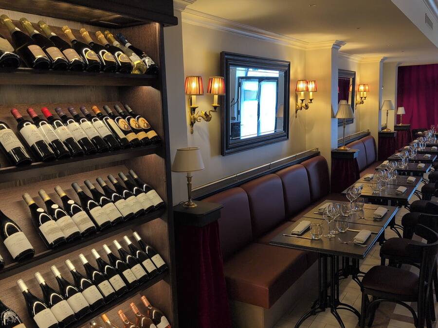 Une salle à manger d’hôtel avec de nombreuses bouteilles de vin exposées sur des étagères, une longue table entourée de chaises et des lampes fixées au mur.