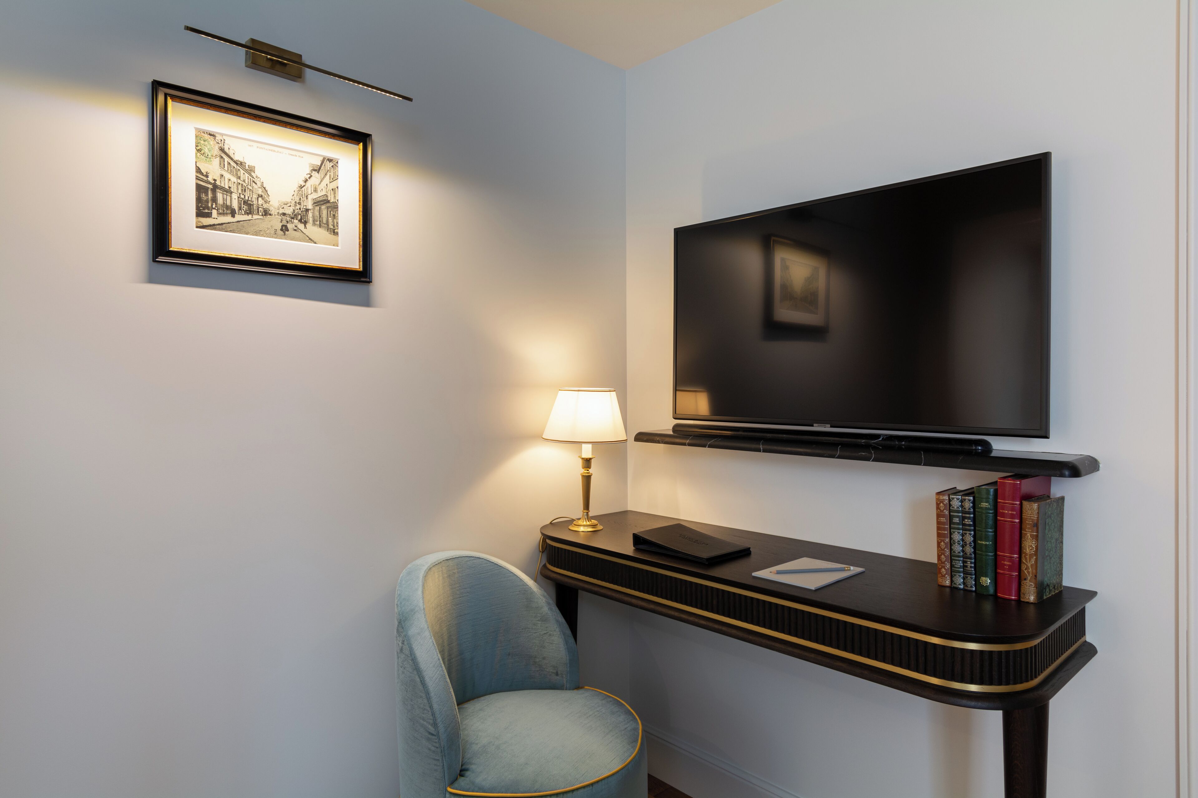 A modern room with a flat-screen TV mounted on the wall, a small table with a lamp, a chair, and a few books.