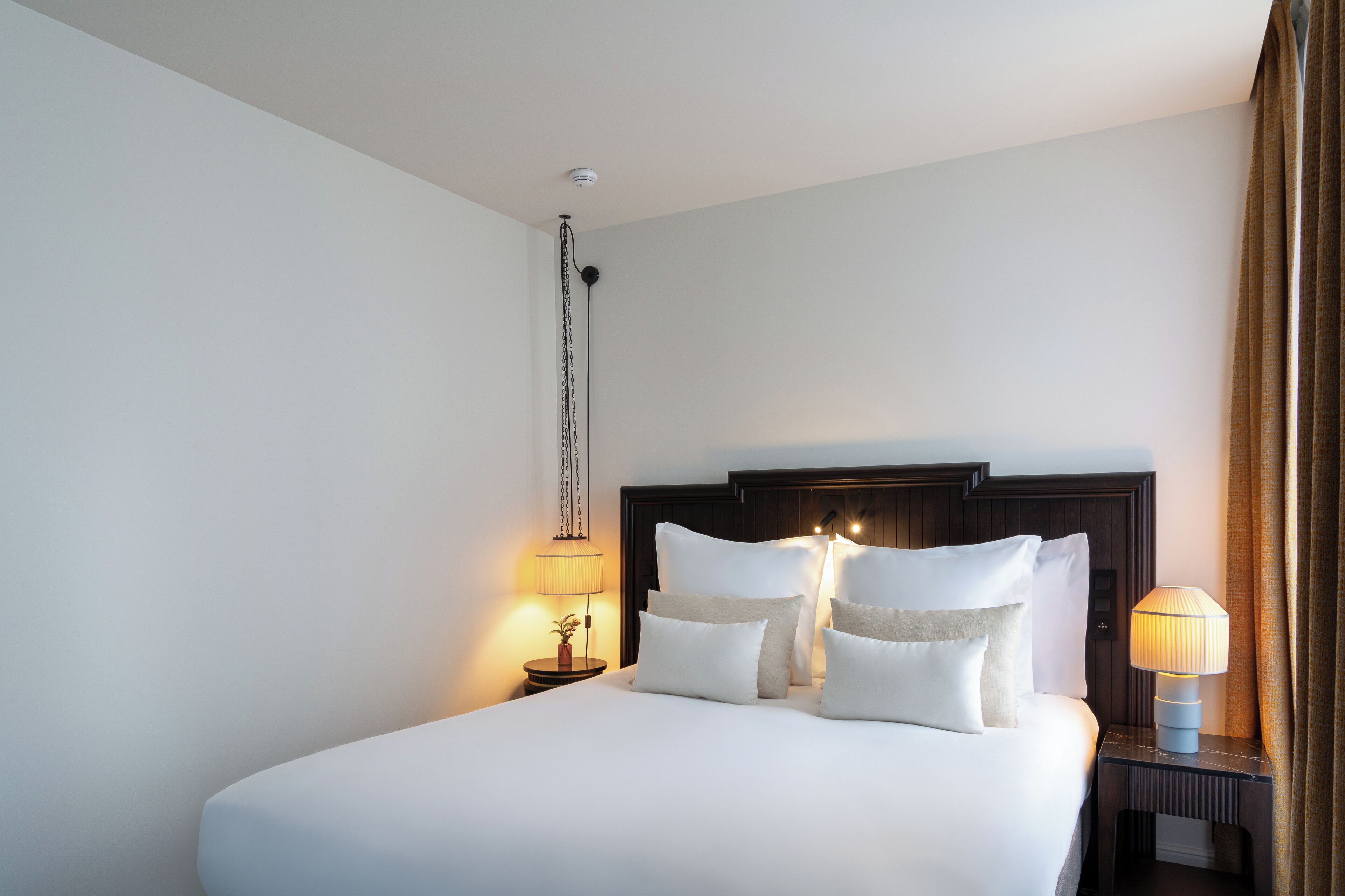 A neatly made bed with white linens and multiple pillows, a dark wooden headboard, a bedside lamp, and a wall-mounted light fixture.