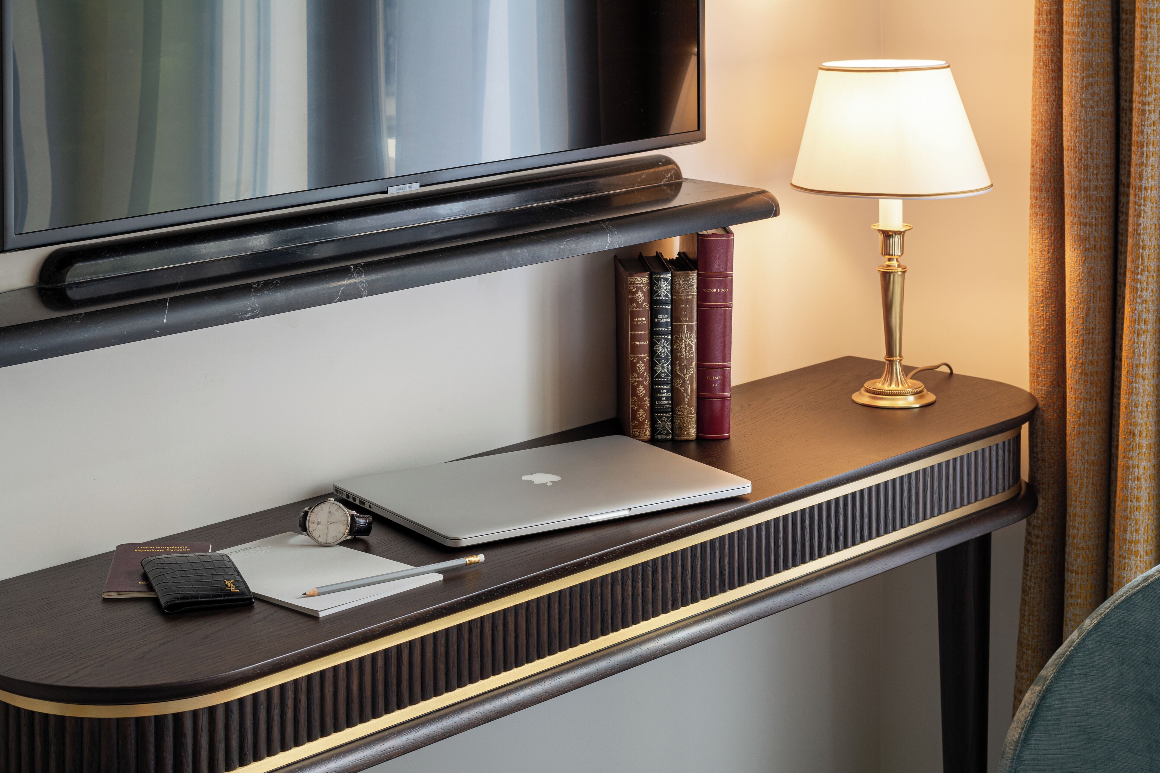 Una scrivania d'hotel ordinata con un portatile, libri, una lampada e un orologio.