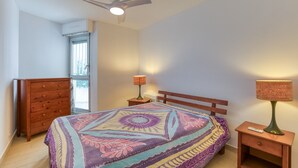 Un dormitorio con cama, cómoda de madera, mesitas de noche, ventilador de techo y una ventana.
