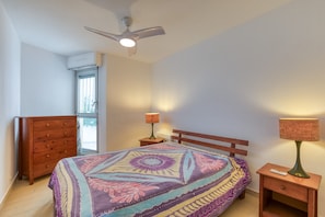 Une chambre à coucher comprenant un lit, une commode en bois, des tables de chevet, un ventilateur de plafond et une fenêtre.