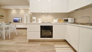 A modern kitchen with white cabinetry, a built-in oven, a microwave, and a coffee maker. There is a dining area with a white table and chairs.