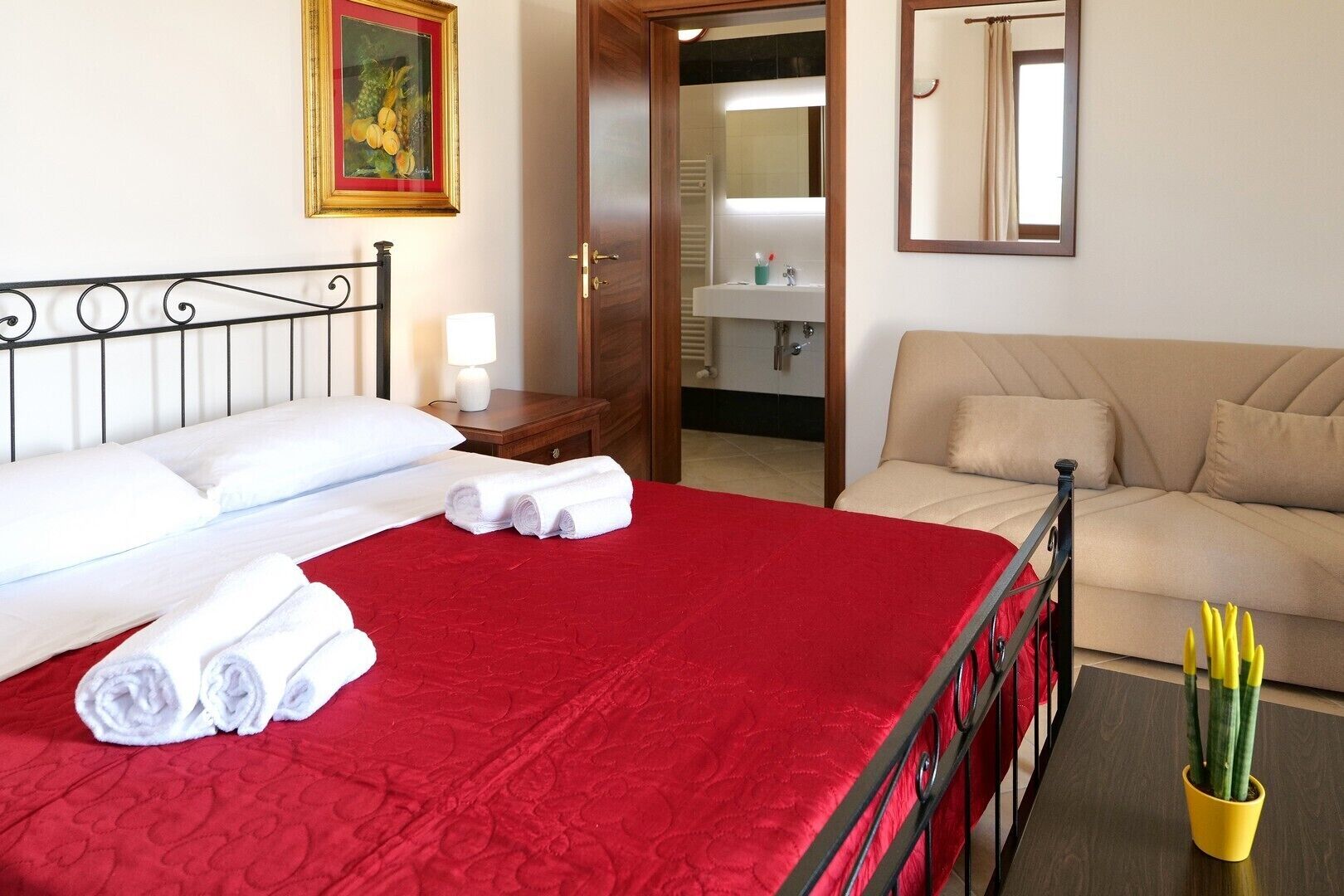 Een hotelkamer met een bed, een rode sprei, een houten nachtkastje, een witte lamp, een beige bank en een kleine potplant.