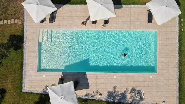 Une vue aérienne d’une piscine rectangulaire à l’eau d’un bleu limpide, entourée d’une terrasse carrelée et de quatre parasols blancs.